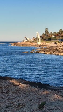 The video shows a seaside view from a rocky cliff. The water is a deep blue with visible ripples on the surface. Across the water, there is a shoreline with buildings and trees visible. A white lifeguard tower stands out against the landscape. The sky is clear and blue. The video is shot vertically.