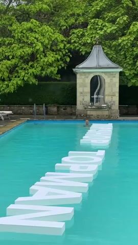 A person walks around the edge of a swimming pool. The pool has large white letters floating on the surface that spell out a word. Another person is swimming in the pool behind the letters. There is a stone structure with a roof behind the pool, and trees with green leaves are visible in the background.