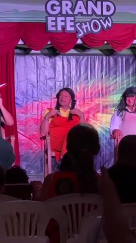 A middle-aged person wearing a bright orange dress and a yellow shirt with braided dark hair, possibly a wig, sits in a light-colored chair on a stage. The backdrop is a shiny, crumpled material with colorful lighting, possibly pink and yellow. Red curtains are visible at the top of the stage, and a sign above reads "GRAND EFE SHOW". In the foreground, the backs of audience members are visible, seated in white chairs. The person on stage gestures with their hands and appears to be speaking.