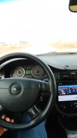 The video shows a view from inside a car, focusing on the dashboard and the road ahead. The car's steering wheel is visible, featuring the Chevrolet logo. The dashboard includes the instrument cluster and a touchscreen displaying a video. The car is driving on a straight, paved road with buildings visible on either side. The sky is bright but somewhat hazy. The interior of the car shows the door panel and side mirror. The person driving is wearing blue jeans.