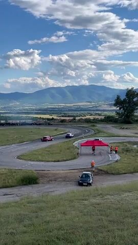 A group of people are gathered near a red tent on a race track. Several cars are on the track, with some appearing to be participating in a race or competition. The track is surrounded by grassy hills and offers a scenic view of mountains and a valley in the background under a partly cloudy sky.