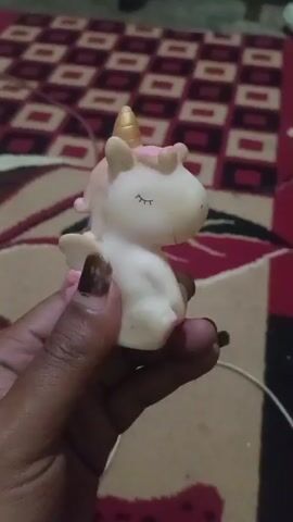 The video shows a hand holding a small, white and pink unicorn toy. The toy has a gold horn and closed eyes. The background appears to be a carpet with a geometric pattern. The hand is holding the unicorn at a close-up angle, and the toy is slightly tilted. The person holding the toy is squeezing it, causing it to make a squeaking sound, as indicated by the timestamps and audio cues. The lighting in the video is dim.
