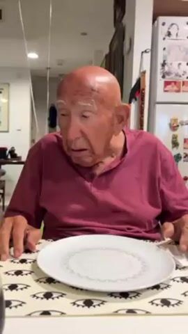 An elderly man with a bald head wearing a maroon shirt sits at a table with a white plate. He speaks and raises his hand in the air. Someone touches his shoulder. The table has a white and black eye pattern table cloth. In the background is a refrigerator covered in photos and magnets.  A balloon with a white string is also visible.
