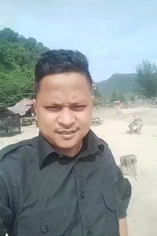 A selfie-style video shows a man with short dark hair and a mustache wearing a black collared shirt. He is standing on a sandy beach with a body of water and waves visible in the background. There are some small huts and trees on the beach. The sky is overcast. The man is looking at the camera and smiling. The video is shaky and appears to be filmed on a mobile device.