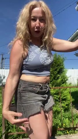 A woman with blonde hair is dancing outdoors on a sunny day. She is wearing a gray crop top and denim shorts. She is in front of a green bush and a white fence. The sky is blue with some clouds.