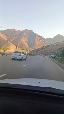 The video shows a rear-facing view from inside a vehicle traveling on a paved road. The road appears to be a highway with multiple lanes and guardrails. The landscape is mountainous, with dry, brown hillsides and clear, blue sky. Several cars are visible on the road, including a white car in front of the viewer's vehicle. There are also signs and utility poles along the side of the road. The overall impression is of a scenic drive through a mountainous region.