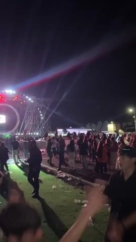 The video begins at a crowded outdoor concert, featuring a stage with complex lighting and a circular screen. The ground is littered with trash, and attendees are visible in the foreground, some dancing or gesturing. The camera moves, capturing a large, blank white screen beside the stage. The video ends abruptly with a blurred shot of a distant cityscape at night, the lights appearing as streaks.