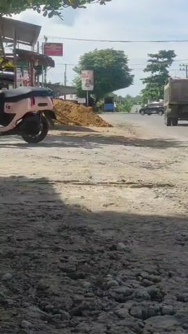 The video shows a street scene with a construction site in the foreground, characterized by dirt and debris. A pink scooter is parked on the left side of the road. In the background, a dump truck is driving on the asphalt road, followed by a motorcycle. Several buildings and trees line the street, under a bright sky.