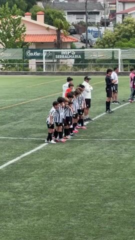 A group of young boys in soccer uniforms stand in a line on a green soccer field. They are facing forward, with their hands clasped in front of them. Some of them are wearing black and white striped jerseys, while others are wearing white jerseys. There are adults standing behind them, some of whom appear to be coaches. A soccer goal is visible in the background, as are buildings and trees. The overall scene appears to be a pre-game ceremony or introduction.