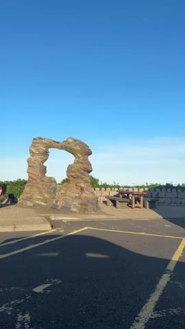 The video shows an outdoor scene on a clear day with a bright blue sky. In the foreground, a paved area with yellow painted lines, possibly a parking lot, is visible. A shadow stretches across the pavement from the right side of the frame. To the left, a large, abstract rock sculpture with an archway is the central focus. The sculpture is made of layered, multi-colored stones. Behind the sculpture and to the right, there is a stone wall or barrier with greenery peeking over it. Further back, a wooden picnic table and benches are situated. The overall environment suggests a public outdoor space, possibly a park or scenic overlook. There are no people visible in the video.