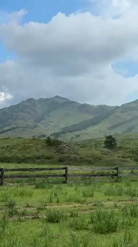 The video shows a wide shot of a pastoral landscape. In the foreground, there is a wooden fence with two horizontal planks and vertical posts. Beyond the fence, a green meadow stretches out, with tall grass in some areas and shorter grass in others. In the middle ground, rolling green hills rise, dotted with trees. A prominent, large mountain range with jagged peaks dominates the background, its slopes covered in green vegetation and showing visible rock formations. The sky above is partly cloudy, with large white cumulus clouds against a bright blue sky. The lighting suggests it is daytime, possibly late morning or early afternoon. No people or animals are visible in the frame. The overall impression is one of a serene and natural environment.