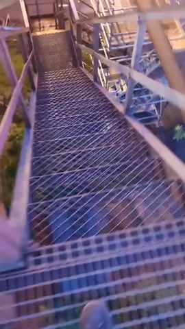 A first-person perspective shot shows a person walking down a metal staircase at night. The staircase has diamond plate treads and is bordered by metal railings. The lighting is dim, with some areas illuminated by artificial lights, casting purple and blue hues. Vegetation and debris are visible to the sides of the staircase. The camera is held at an angle, facing downwards along the stairs.