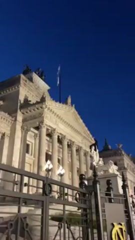 The video shows a building in Argentina at night. The building is white and has a classical design with columns and statues. An Argentinian flag flies from the top of the building. The sky is dark blue. A black wrought iron fence runs along the front of the building with an emblem attached to one of the gate posts.