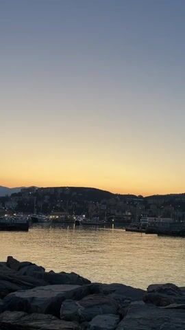 This is a vertical video capturing a sunset scene over a harbor in Imperia, Italy. The foreground features a cluster of large, rough-hewn rocks, suggesting a breakwater or rocky shoreline. Beyond the rocks, the calm water of the harbor reflects the warm hues of the setting sun, creating a shimmering golden surface.

In the middle ground, a marina is visible with several boats and yachts docked. Lights are starting to twinkle from buildings that line the harbor and extend up the gentle, tree-covered hills in the background. The overall impression is one of a peaceful evening in a coastal town.

The sky transitions from a deep blue at the top of the frame to a soft, warm orange-yellow band just above the horizon, characteristic of sunset. The lighting is dim, indicating it is either dusk or early evening. The weather appears to be clear and calm, with no strong winds or clouds visible. The scene evokes a sense of tranquility and beauty, typical of a seaside sunset. There is no visible text in the video.
