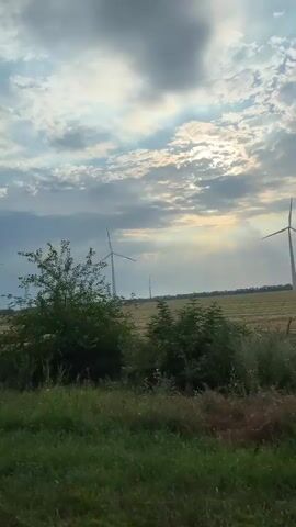 A video captured from a moving vehicle shows a rural landscape under a cloudy sky. The sun is visible through breaks in the clouds, casting light and creating a slight haze effect. In the foreground, there is green grass and some bushes. In the middle ground, a field can be seen, and further in the distance, there are several wind turbines spread across the field. The vehicle appears to be driving along a road adjacent to the field.