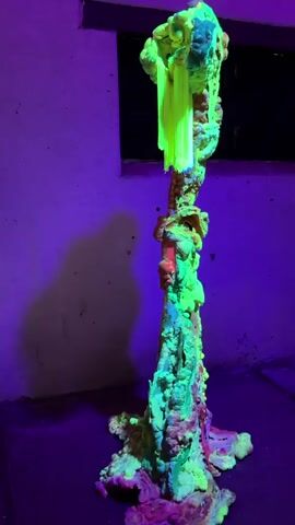 A neon green, textured sculpture resembling a twisted tree trunk stands on a dark surface under purple lighting. The sculpture has pink and red accents at its base. A fire extinguisher is visible on the wall to the left of the sculpture.