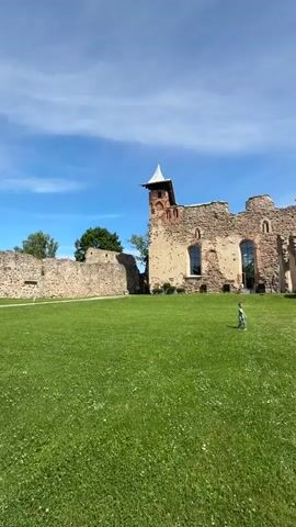 A lone child is seen walking across a lush green lawn in front of the ruins of an old stone castle under a clear blue sky with scattered clouds. The castle is partially intact, with crumbling walls and arched openings. The child appears to be walking away from the camera. The sun is shining, casting shadows on the grass. The scene suggests a historical landmark being visited on a pleasant day.