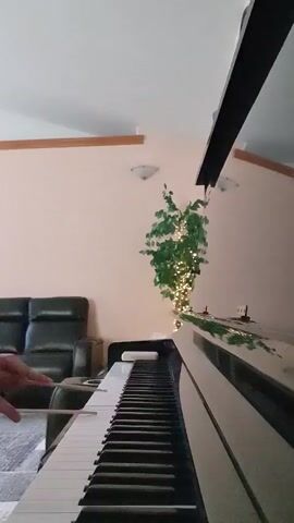 A vertical shot captures a person's hands playing a piano, with drumsticks in their hands. The piano is glossy black and white, and is positioned in the foreground. In the background, there is a black leather sofa, a potted plant decorated with string lights, and a pale pink wall. The ceiling has a sloped section with exposed beams.