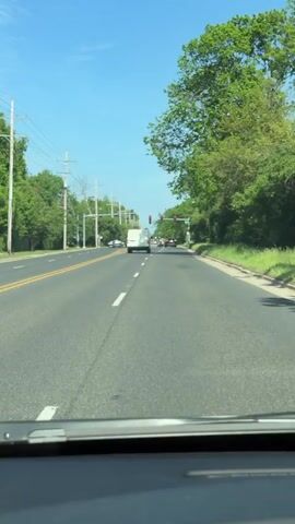 The video depicts a view from inside a car driving on a road. The road has two lanes in the direction of the view, separated by double yellow lines. A white line marks the right edge of the lane. On either side of the road are green trees and vegetation. Overhead, there are utility poles and wires stretching along the road. In the distance, there's a traffic light showing both red and green lights. Several vehicles are ahead, including a white van. The sky is blue with a few scattered clouds. The dashboard of the car is visible at the bottom of the frame.