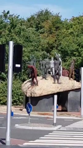 This is a medium shot video taken from a low angle on a sunny day. It shows a sculpture of several human figures atop a large rock formation. The figures are depicted in various poses, some standing and some crouching. The sculpture is located near a pedestrian crossing with white stripes on the road. In the foreground, there are two silver poles with traffic lights attached. A blue circular sign with a white arrow pointing left is visible on a pole in front of the sculpture. Behind the sculpture, there is a dense wall of green trees. The overall environment appears to be an outdoor public space, possibly a park or a street intersection.