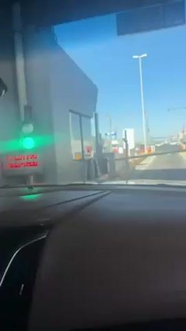A car is at a toll booth, ready to proceed through as indicated by the green light. The toll booth has a traffic light, a display that reads "IRAN VIAL", and a barrier arm raised. The environment suggests a clear, bright day with a blue sky. Other toll booths are visible in the distance, and the car's dashboard is visible in the foreground.