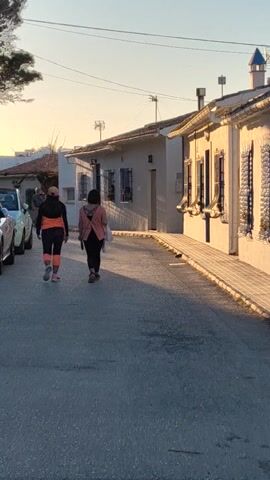 The video shows a low-angle, steady shot of two people walking down a narrow street, with a white building on the left side and parked cars partially visible at the bottom left. The first person is wearing a black shirt, black pants, and a pink jacket. The second person is wearing an orange shirt, black pants with orange stripes, and a hat. The building has windows with metal bars and a simple facade. The sky is clear and light blue. The street appears to be made of asphalt, with some shadows cast by the building.