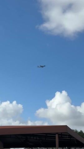 The video shows an airplane flying in the sky. The sky is blue with fluffy white clouds. In the foreground, there's a brown rooftop and some trees. The plane is high in the sky, appearing as a small silhouette against the blue background. The weather seems pleasant with clear visibility.