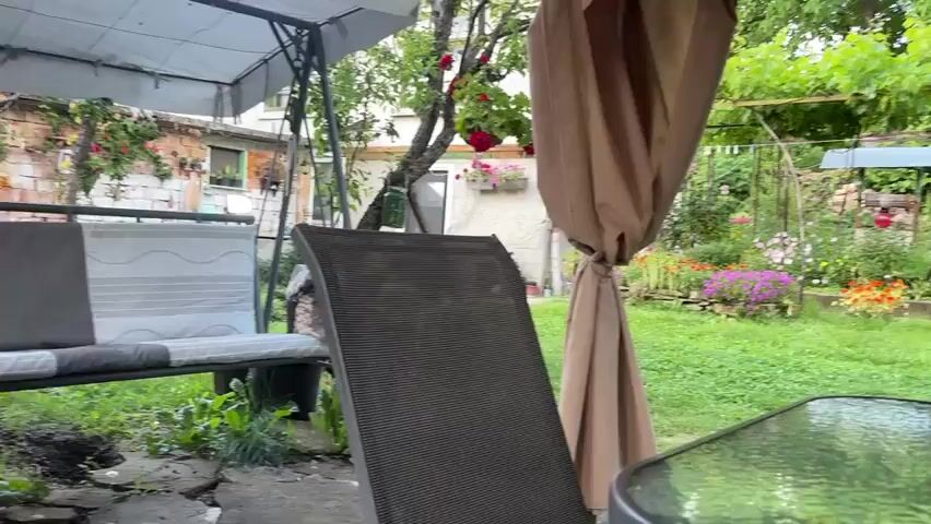 The video shows a backyard scene with a glass-topped table in the foreground. On the table, there is a pack of cigarettes, an ashtray, and a clear plastic container. Two chairs are visible behind the table, and a tan gazebo covers part of the area. In the background, there is a lush garden with various flowers, a brick wall, a wooden ladder leaning against a structure, and trees with vines growing on them. The overall impression is of a relaxed outdoor setting.