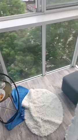 The video begins by showing a hookah on a small, round table. A blue towel is draped over the table, and a white fluffy rug is next to it. The view then pans to a window overlooking a town with trees and mountains in the distance. The town appears to be in a valley, with some buildings and a factory visible. The mountains are large and majestic, with some clouds around their peaks. The sky is a clear blue.