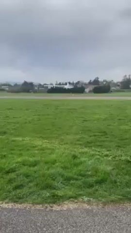 The video shows a white helicopter taking off from a flat, grassy field. In the background, there are trees, buildings, and a cloudy sky. The helicopter rises from the ground and begins to move away from the camera. The sound of the helicopter's engine is audible.