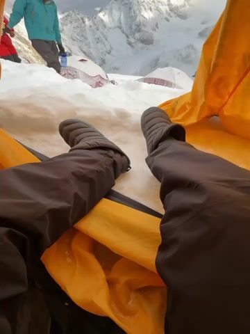 The video shows a first-person perspective from inside a tent with the flap open. The viewer's legs, clad in puffy slippers and dark pants, are visible in the foreground. Outside the tent, a snowy, mountainous landscape unfolds under a cloudy sky. Two individuals are visible in the mid-ground. One person, wearing a bright red and black suit, is holding hands with another person dressed in a teal jacket and dark pants, who is reaching for a blue water bottle. Several other tents, some with red coverings, are scattered in the snowy terrain. The mountain peaks in the background are snow-covered and dramatic. The scene appears to be a high-altitude base camp or a similar mountain environment.