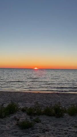 The video captures a tranquil beach scene at sunset. The sun, a small, vibrant orange orb, is setting just above the horizon line of the vast ocean. Its reflection creates a shimmering path of light across the rippling water. The sky above is a gradient of soft blues and oranges, transitioning from clear blue at the top to a warm, fiery orange closer to the horizon. The foreground consists of a sandy beach with a few small, green plants growing on it. The waves gently lap at the shore, creating a peaceful and serene atmosphere. The overall impression is one of natural beauty and tranquility as the day comes to a close.