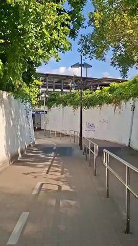 The video shows a paved walkway leading to an underground tunnel. The walkway has white dashed lines painted on it, indicating a bike lane. There are metal railings on the right side of the walkway. The tunnel entrance is visible in the distance, and a sign above the entrance indicates "S 1-7" and "Willkommen am Bahnhof Baden". The tunnel walls are covered in white graffiti. Trees and foliage are visible on the left and right sides of the walkway, and a street lamp is positioned near the tunnel entrance. The sky is blue with some clouds. The video appears to be filmed from a moving camera, possibly a person walking.