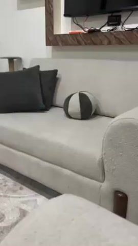 The video shows a close-up of a living room setting. A light beige couch is the main focus, adorned with two dark grey square pillows and a round pillow with alternating grey and beige sections. A television mounted on the wall with a brown wooden frame is visible in the background along with power cords and a cable box. Part of a patterned rug is visible in the lower left corner.