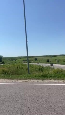 The video shows a sunny, clear day with a bright blue sky. A paved road with a white line marking the edge curves into the distance. On the left side of the road, there are green fields with some bushes and trees. A tall pole with a solar panel on top stands near the side of the road. The camera appears to be moving, possibly from a vehicle, as the landscape shifts slightly. In the distance, the fields continue to roll and meet the horizon.
