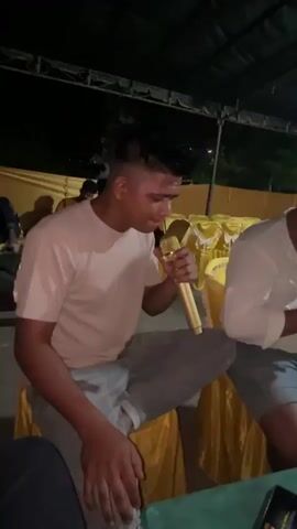A young man with short, spiky dark hair is sitting on a chair with a gold microphone in his hand, which he holds close to his mouth. He is wearing a light pink t-shirt and gray pants. He is looking down and appears to be singing or speaking into the microphone. To his right, another person in a white shirt is partially visible. The background is dark and appears to be an outdoor setting at night, possibly under a tent or canopy, with yellow fabric draped around the area. There are other people and objects in the background, but they are out of focus. The overall lighting is dim.
