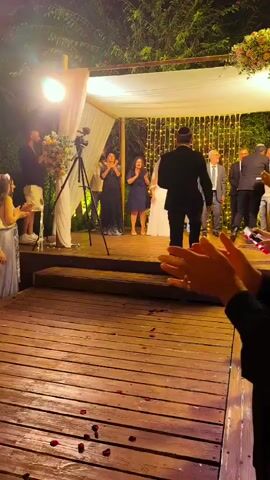 The video shows an outdoor evening wedding ceremony. In the background, a bride in a white dress and groom in a black suit are standing under a chuppah, a canopy. Several people are standing on a raised wooden platform behind them, including two women in dark blue dresses, two men in suits, and others whose faces are not clearly visible. A camera on a tripod is set up on the platform. In the foreground, the lower half of several people's bodies are visible, and they are clapping. The ground in front of the platform is made of wood and is scattered with rose petals. The overall atmosphere appears festive and celebratory.