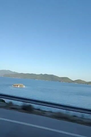 The video shows a view from a vehicle moving alongside a body of water. The water is blue and appears calm, with a shoreline and hills in the distance. The sky is clear and blue. The foreground is dominated by a guardrail and the side of the road, with trees passing by. The camera seems to be positioned to capture the scenery as the vehicle travels. There are no people visible in the video.