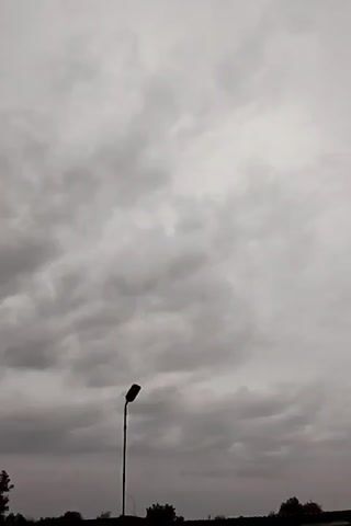 This is a vertical video recording of a cloudy sky with a lamppost in silhouette against it. The sky is filled with various shades of gray clouds, suggesting an overcast or potentially stormy day. The lamppost is a tall, slender object with a distinct curved top, casting a dark shadow. The lower portion of the frame shows a dark, silhouetted line of trees and possibly the horizon, indicating an outdoor setting. There are no people visible. The overall mood is somber due to the cloudy sky. The location context suggests this scene is from the village of Kiyaly in Kazakhstan. The time of day is ambiguous due to the overcast conditions, but the lack of bright light suggests it is likely not midday. No text is visible in the video.