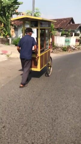 A man in a dark blue shirt, dark pants, and a black cap pushes a yellow and brown food cart down a paved road. The cart has a sign that reads "BAKSO". The man's left hand is on the cart's handle, and his right hand is holding it from the side. He is walking away from the camera. The road is asphalt, and there is vegetation on the sides, including trees and bushes. A building with a tiled roof is visible in the background. The sky is clear and appears to be daytime.