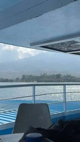 A person's hand places a blue-capped plastic water bottle on a table. A portion of a white chair is visible. The view is from the deck of a ferry looking out at a body of water with a distant mountain range. The sky is overcast with clouds. The deck of the ferry has blue and white striped flooring and a metal railing.