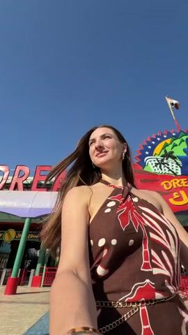 The video begins with a medium shot of a woman with long, dark hair, wearing a patterned halter top and large hoop earrings. She is standing in front of a large, colorful sign that reads "DREAM PARK" in large red letters. The sign is topped with a cartoonish depiction of mountains and palm trees, and a flag waves from a pole above it. The woman is looking slightly up and to her right, with a slight smile on her face. Her right arm is extended towards the camera, as if she is taking a selfie. The background appears to be an amusement park or entertainment venue, with pathways and structures visible. The sky is a clear blue. The woman then turns her head slightly to the left and puckers her lips. The scene continues with more close-ups of the woman's face as she moves slightly and her hair blows in the wind. The sign and park entrance remain in the background.