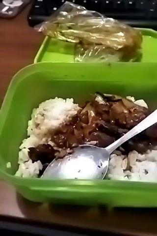 A close-up shot of a green plastic lunch container filled with white rice and what appears to be dark meat covered in a glossy sauce, possibly beef or chicken. A silver spoon is resting in the food. In the background, another green container is visible with some food wrapped in clear plastic. The setting appears to be a desk or table, with a black keyboard visible behind the lunch containers. The lighting is dim, suggesting an indoor environment. The camera movement is minimal, with a slight zoom in over the course of the video.