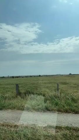 The video begins with a view of a green grassy field with a dirt path in the foreground and trees and a cloudy sky in the distance. The camera pans to the right, showing a car's side mirror and then moves inside the car, looking through the windshield. The car is on a paved road with other vehicles in the distance. The environment appears to be rural with fields and trees on either side of the road. The sky is blue with some clouds. The interior of the car shows a dashboard and a windshield.