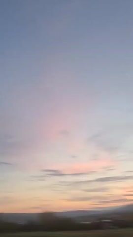 The video shows a landscape at dusk with mountains in the distance. The sky is transitioning from a pale blue to shades of orange and pink. There are some scattered clouds. In the foreground, there is a dark, blurry field. There are no people or specific objects clearly visible. The camera appears to be moving slightly.