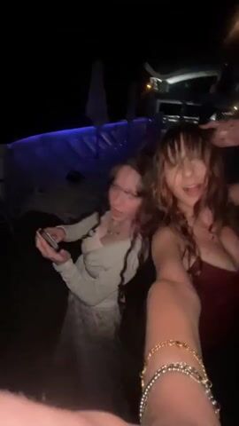 Two young women are dancing and filming a video at night. The woman on the left is wearing a light blue cardigan over a white top and a patterned skirt. She is holding a phone and has her hair down. The woman on the right is wearing a dark red dress and has her hair down. She is smiling and her mouth is open. They are outdoors, with blue lighting and some umbrellas in the background.