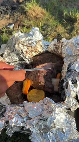 A close-up shot shows a person's hands using a knife to cut into a piece of meat wrapped in aluminum foil. The meat appears to be cooked, with some parts showing a reddish-pink interior. Slices of lemon and orange are visible around the meat, as well as some root vegetables. The foil is slightly crumpled, and some dirt and debris are present on the meat and the foil. The background is out of focus, but appears to be an outdoor setting with grass and a hint of smoke or ash, suggesting the meat may have been cooked over a fire. The lighting suggests it is daytime, possibly late afternoon given the warm tones.