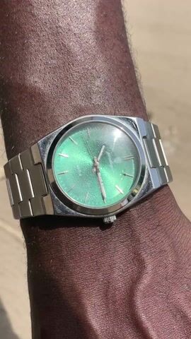 A close-up shot shows a dark-skinned hand wearing a silver watch with a green face. The hand is resting on an object that appears to be part of a bag or box, featuring a green camouflage pattern and the words "RINGTEA GOODME" printed on it. A black selfie stick with a small ring light attached is also present, along with a green baseball cap that has the logo of the Chicago White Sox and "WORLD SERIES CHAMPIONS" embroidered on it. The finger briefly presses the button on the selfie stick.