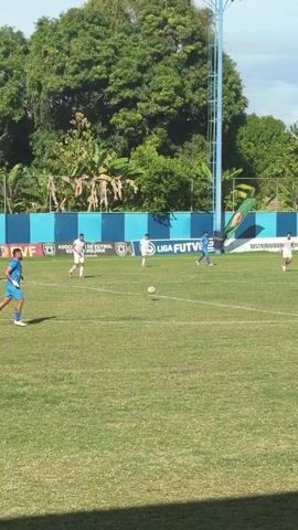A wide shot shows a soccer match in progress on a sunny day. The camera is positioned on the side of the field, looking across the pitch towards the other side. Several players in light blue and white uniforms are on the field. The player in light blue with the number 5 is dribbling the ball on the left side of the frame, moving towards the center. Another light blue player in the background on the far right of the frame also has the ball. The field is green and well-maintained. Behind the field, there's a blue and white striped wall, trees, and some buildings. The sky is partly cloudy.