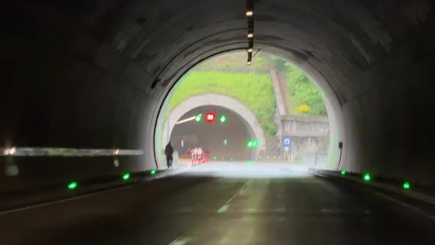 The video shows a view from inside a tunnel looking out at the exit and the road beyond. The road is wet and there are green lights lining the edges. Outside the tunnel exit, there is another tunnel entrance, a grassy slope, road signs, and traffic cones. A person walks along the side of the road near the tunnel exit. The signs above the second tunnel entrance show an 80 speed limit. The video appears to be taken from a moving vehicle.